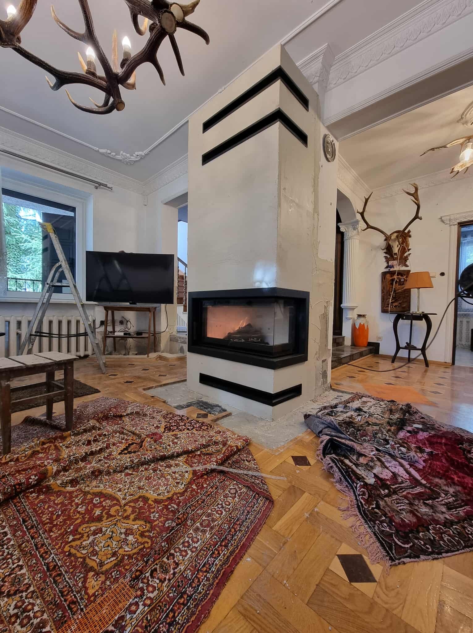 Wnętrze w trakcie prac: kominek z czarnymi akcentami, zabezpieczony dywanami, drabina oparta o ścianę. Widoczne ślady remontu na podłodze i ścianach, elementy dekoracyjne.