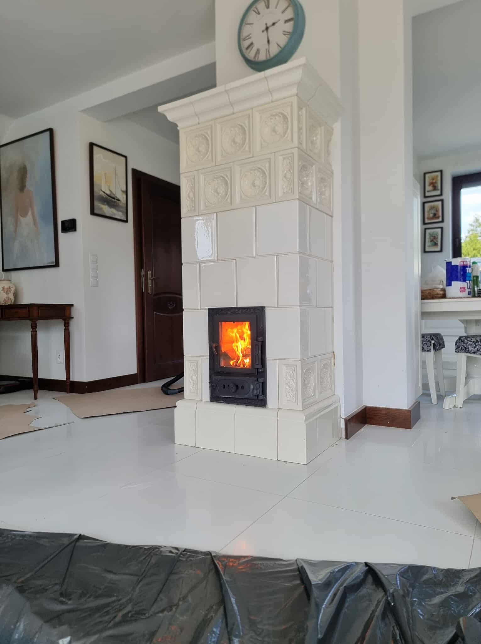 Biały, kaflowy piec w salonie z palącym się ogniem w palenisku. Widoczne elementy wystroju wnętrza: zegar, obrazy, stół i krzesła. Podłoga zabezpieczona folią.