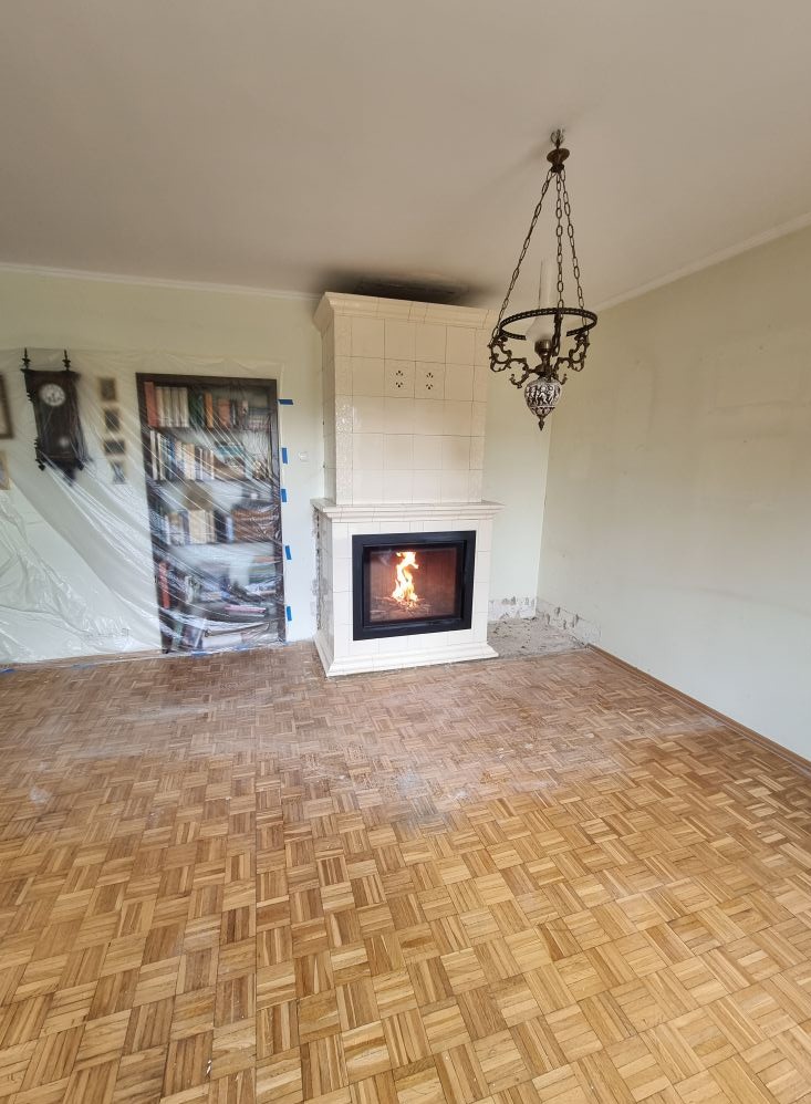 Biały, kaflowy kominek z paleniskiem w salonie w trakcie wykańczania. Widoczny parkiet i lampa w stylu retro. Usługi zduna w Kolsku.