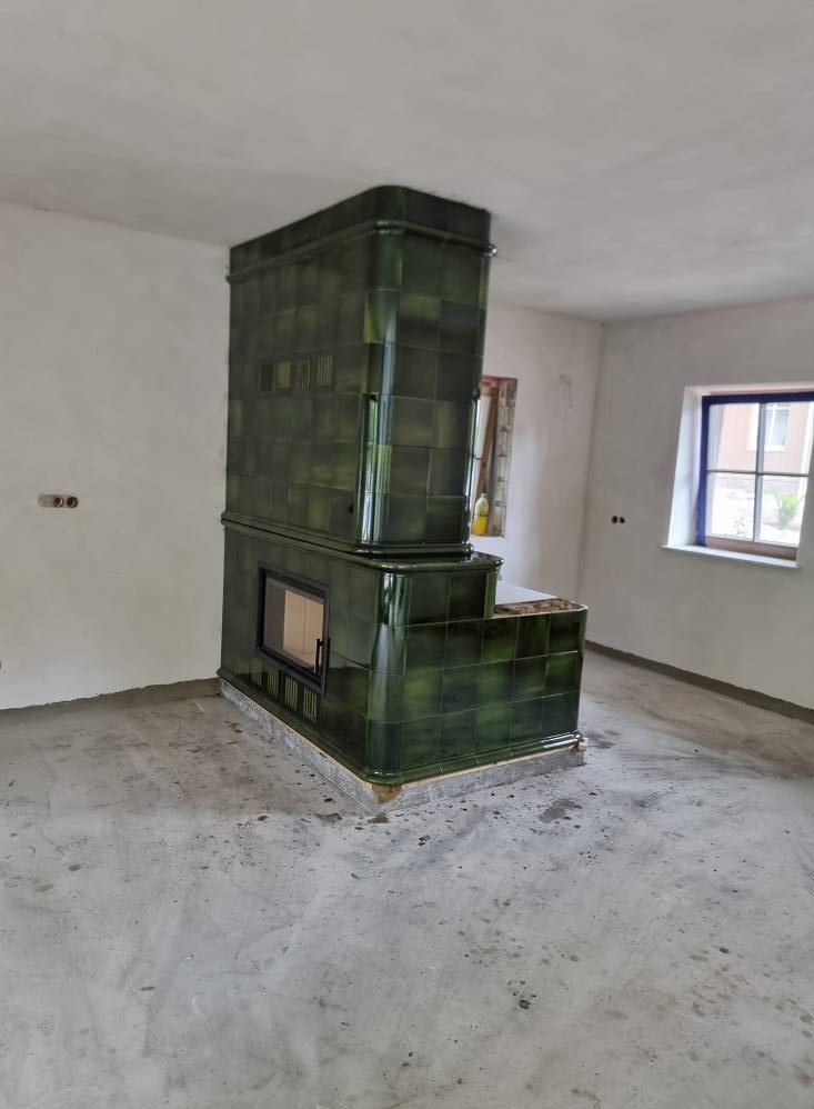 Zielony piec kaflowy w surowym wnętrzu z oknem. Widoczne niedoskonałości ścian i podłogi podkreślają ręczne wykonanie i tradycyjny charakter.