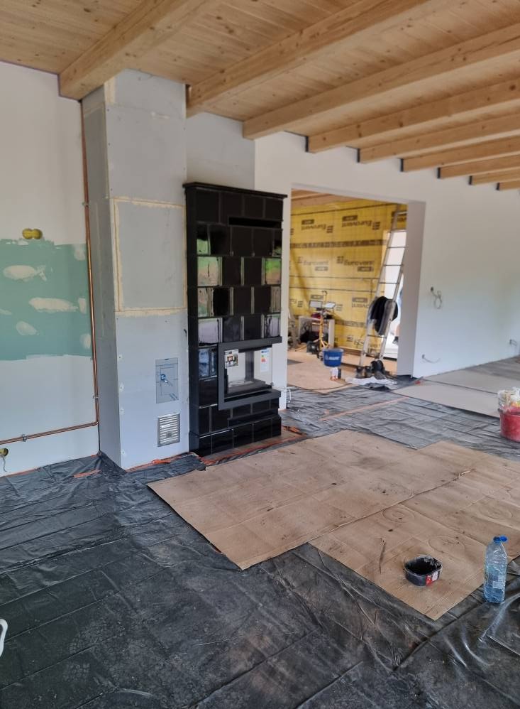 Czarny, kaflowy piec w surowym wnętrzu. Podłoga zabezpieczona folią, widoczne drewniane belki stropowe. Piec w trakcie montażu lub tuż po nim.