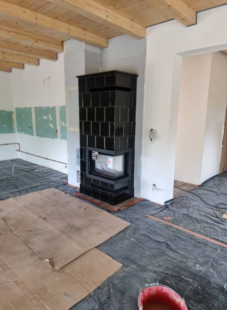 Czarny, kafelkowy kominek narożny w surowym wnętrzu. Widoczne zabezpieczenia podłogi i drewniany strop. Kątowe ujęcie, prezentujące detale wykończenia.