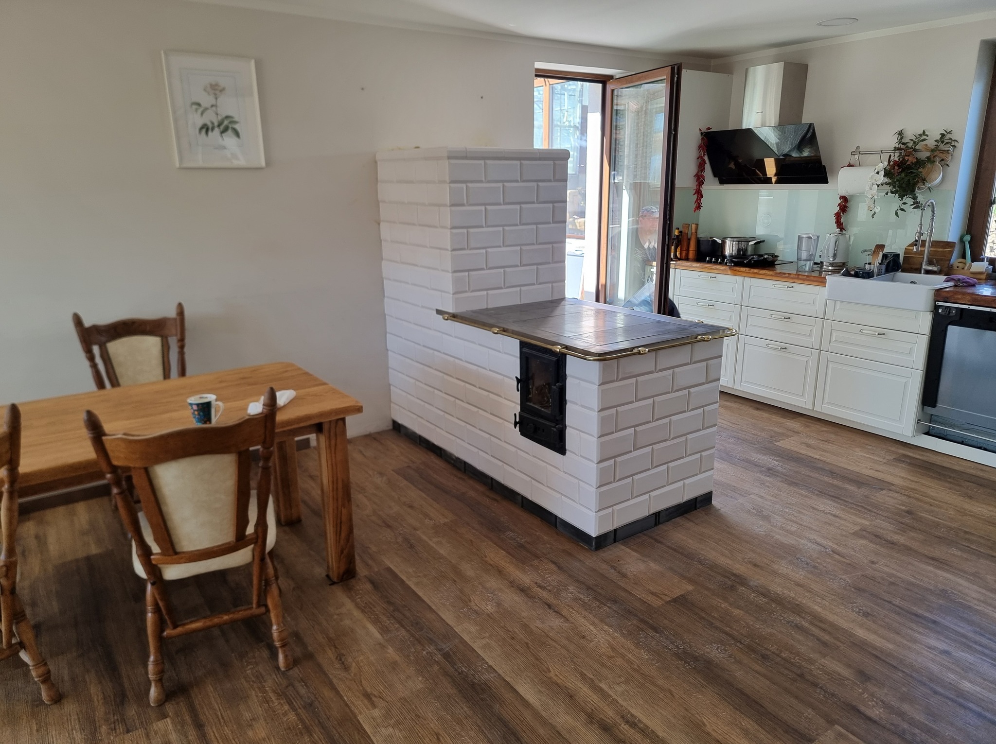 Biały, kaflowy piec kuchenny w rustykalnym wnętrzu z drewnianą podłogą i stołem. Widok na otwartą kuchnię z jasnymi szafkami i oknem.