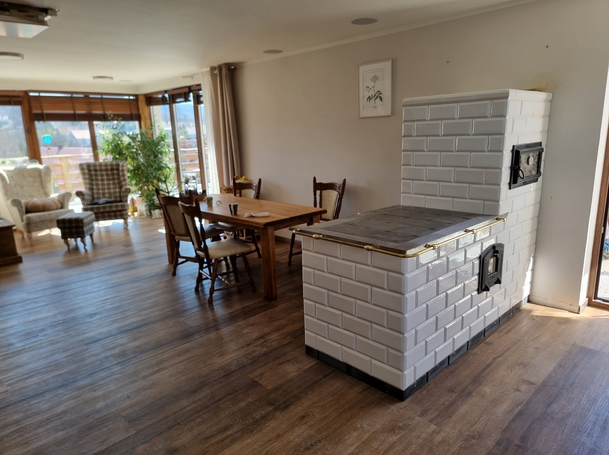 Biały piec kaflowy w salonie z drewnianym stołem i krzesłami. Jasne wnętrze z parkietem i dużymi oknami. Widoczny fragment pieca z metalowymi drzwiczkami.