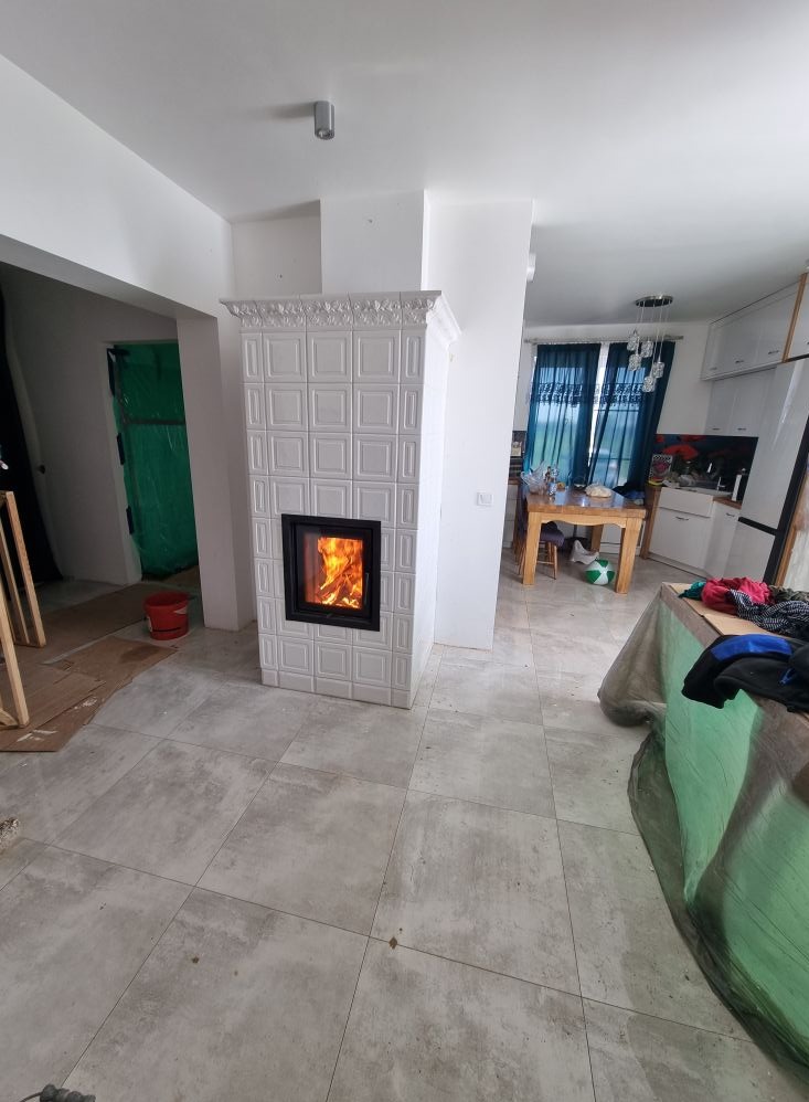 Biały, kaflowy piec kominkowy z paleniskiem, w trakcie wykańczania wnętrza. Widoczne płytki podłogowe i elementy kuchni w tle. Otwarta przestrzeń dzienna.