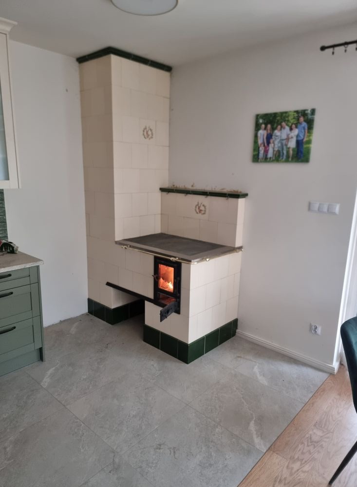 Tradycyjny piec kaflowy w kuchni, z palącym się ogniem w palenisku. Białe kafle z zielonymi akcentami. Na ścianie wisi rodzinne zdjęcie. Szare płytki na podłodze.