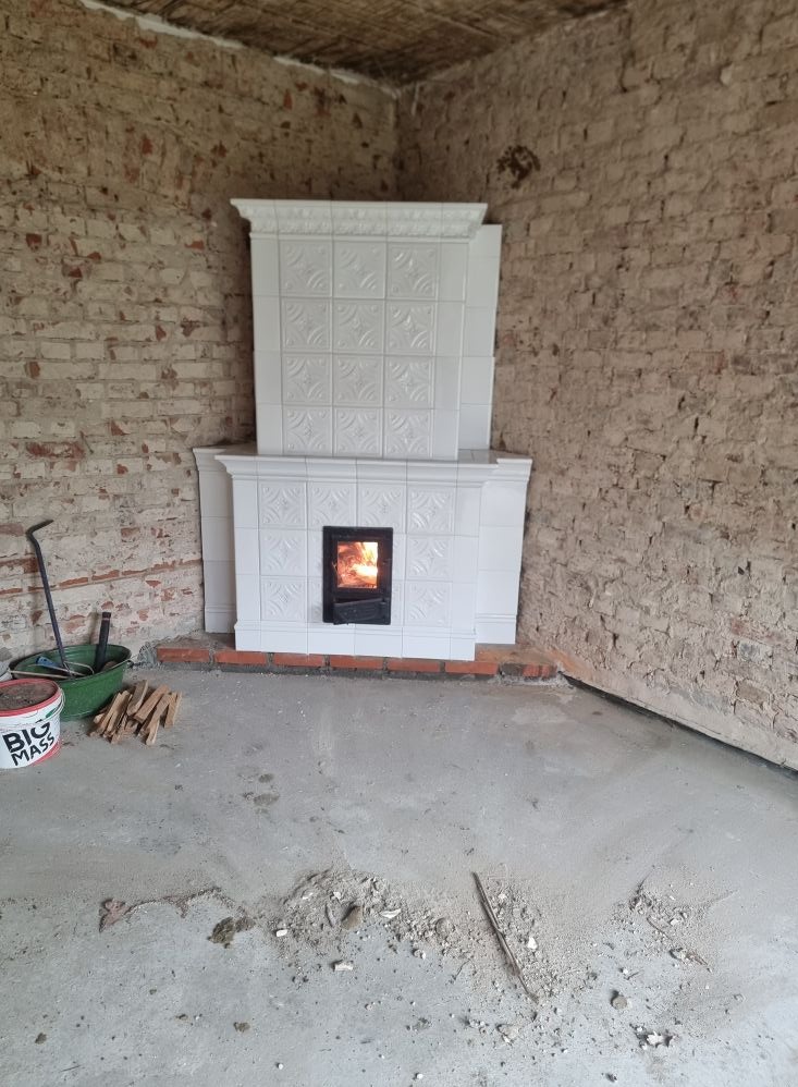 Biały piec kaflowy z palącym się ogniem w surowym wnętrzu z ceglanymi ścianami. Na podłodze widoczne ślady budowy i narzędzia.