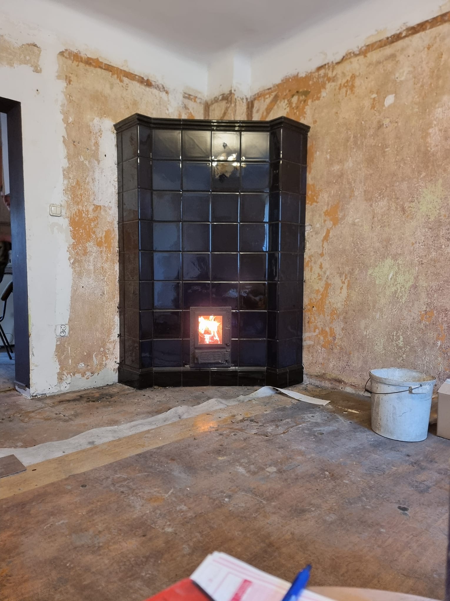 Czarny, kaflowy kominek narożny z palącym się ogniem w surowym wnętrzu z fragmentami odrapanych ścian i przygotowanym podłożem. Widoczne wiadro i przybory.