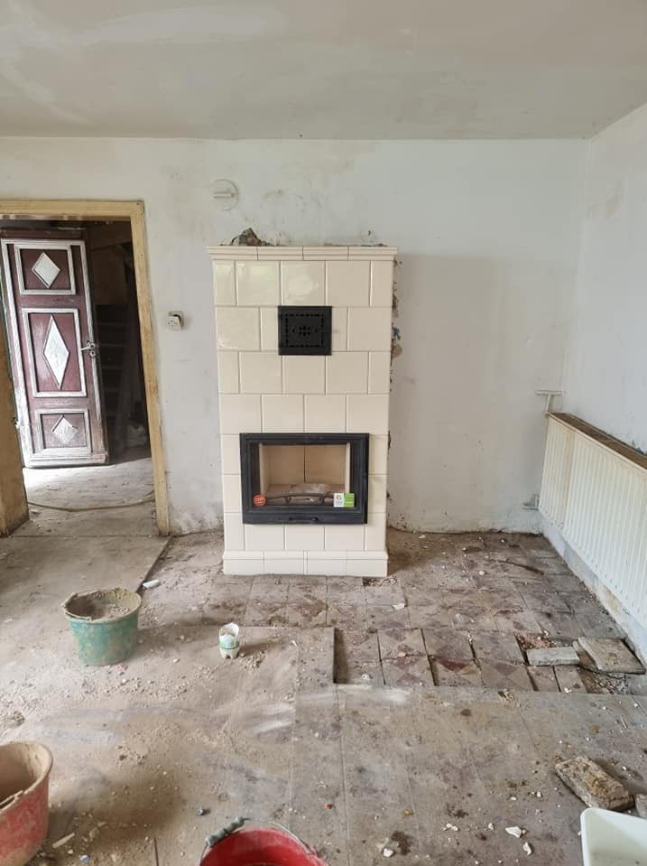 Wykończony, biały, kaflowy kominek z czarnym wkładem w remontowanym pomieszczeniu. Widoczne ślady prac i przygotowania do dalszych etapów wykończeniowych.