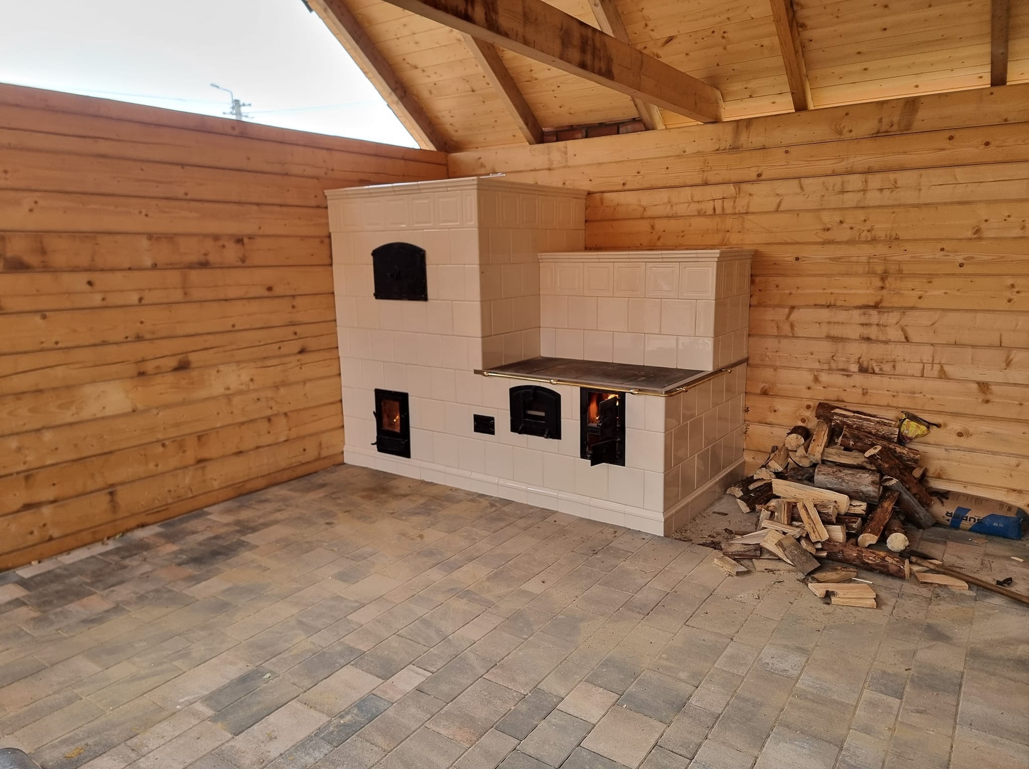 Biały, kaflowy piec z paleniskiem w drewnianej altanie. Widoczne płomienie, ułożone drewno obok. Kafelki z delikatnym wzorem. Podłoga wyłożona kostką.