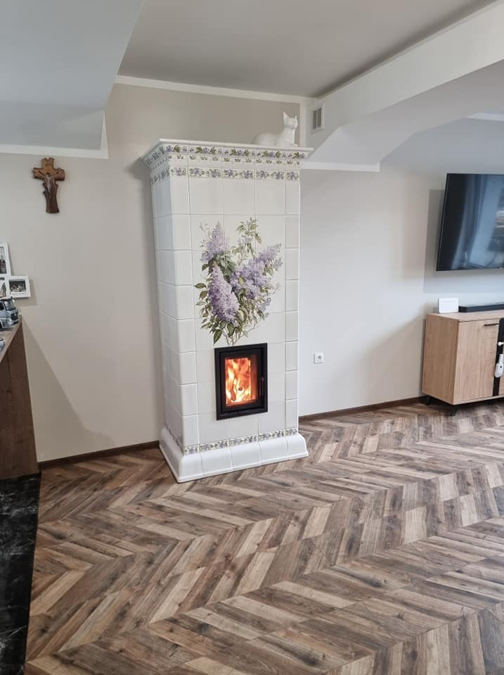 Biały, kaflowy piec z motywem bzu w salonie, z paleniskiem i dekoracyjną figurką kota na szczycie. Parkiet ułożony w jodełkę.