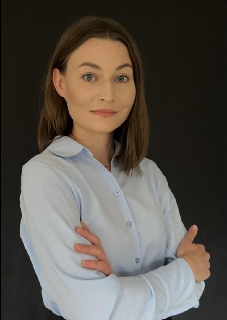 Portret kobiety w błękitnej koszuli na ciemnym tle, skrzyżowane ręce, stonowany makijaż, profesjonalny wygląd.