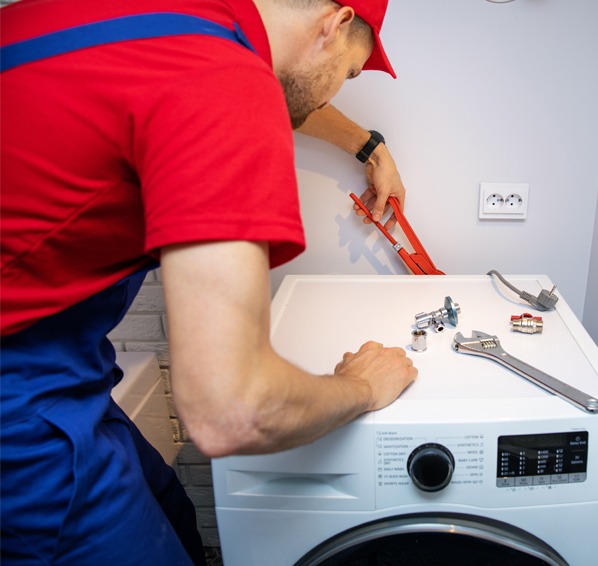 Serwisant w czerwonej czapce naprawia pralkę, używając klucza i narzędzi. Na pralce leżą elementy instalacji wodnej. Biała ściana z gniazdkiem w tle.