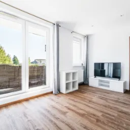 Jasne, przestronne wnętrze z parkietem, balkonem i telewizorem. Minimalistyczny design, białe ściany, drewniane akcenty i naturalne światło. Widok na zieleń z balkonu.