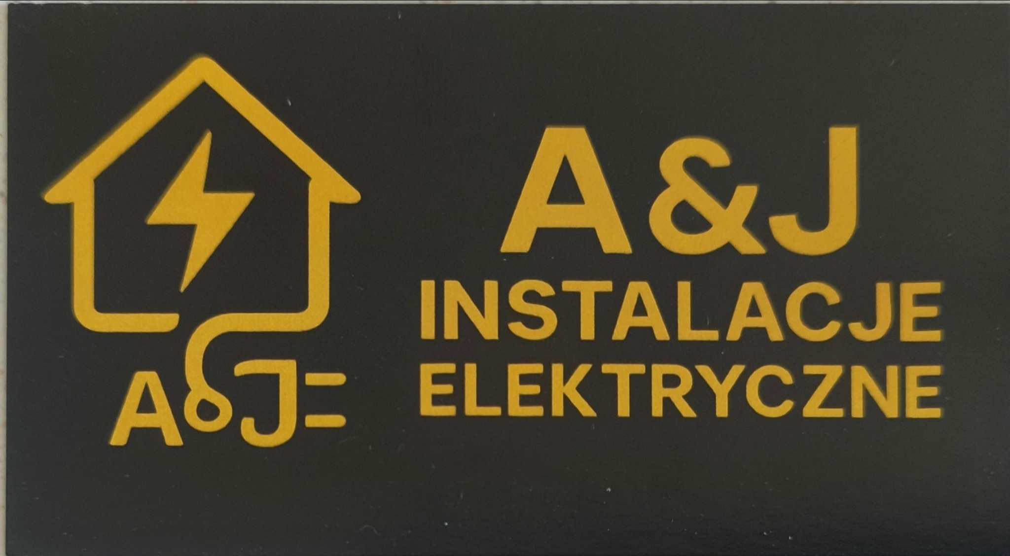 Żółte logo firmy A&J Instalacje Elektryczne na czarnym tle. Symbol domu z błyskawicą i napis 'INSTALACJE ELEKTRYCZNE'.