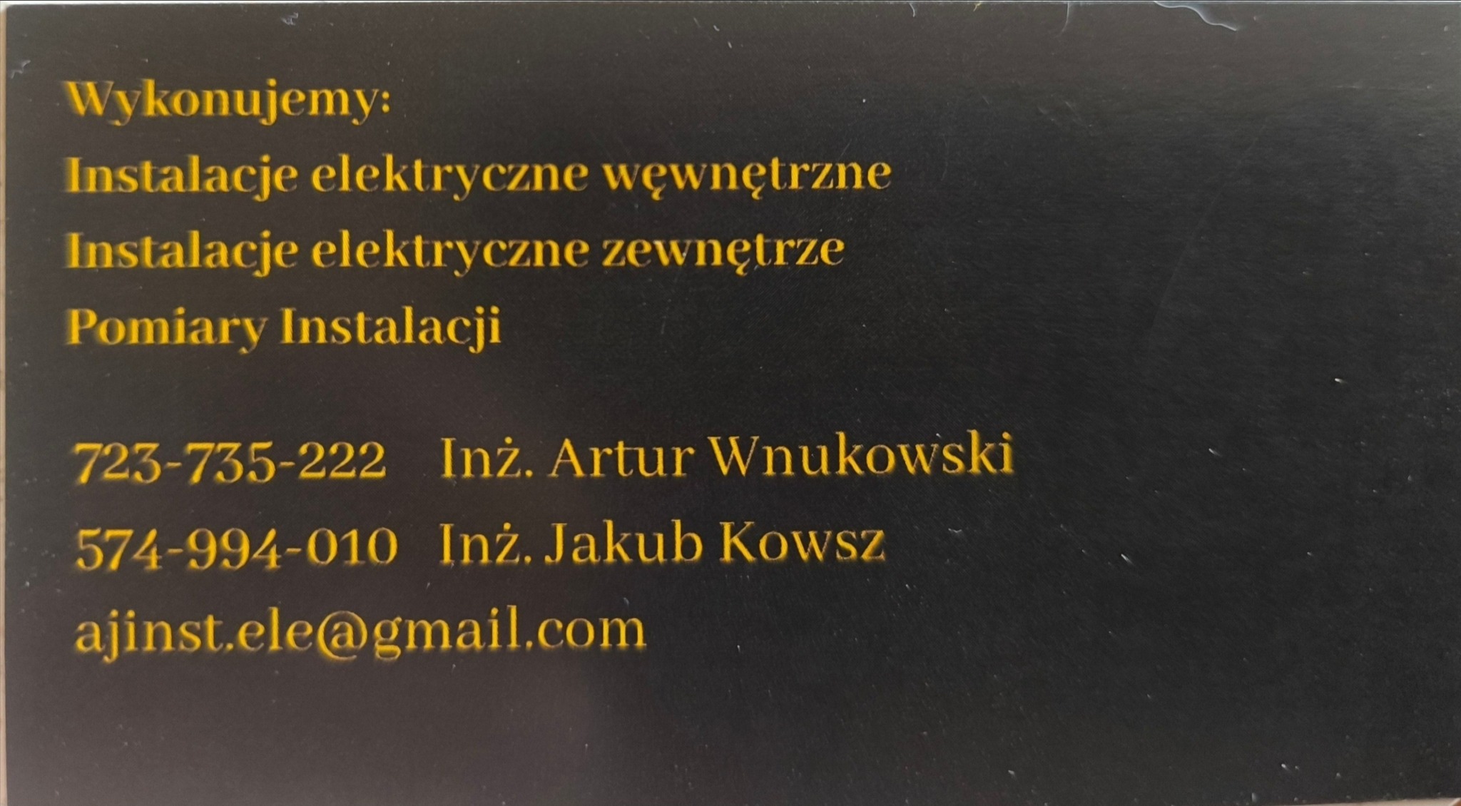 Wizytówka firmy elektrycznej oferującej instalacje wewnętrzne, zewnętrzne i pomiary. Dane kontaktowe: numery telefonów, adres e-mail. Czarny kartonik, złote litery.