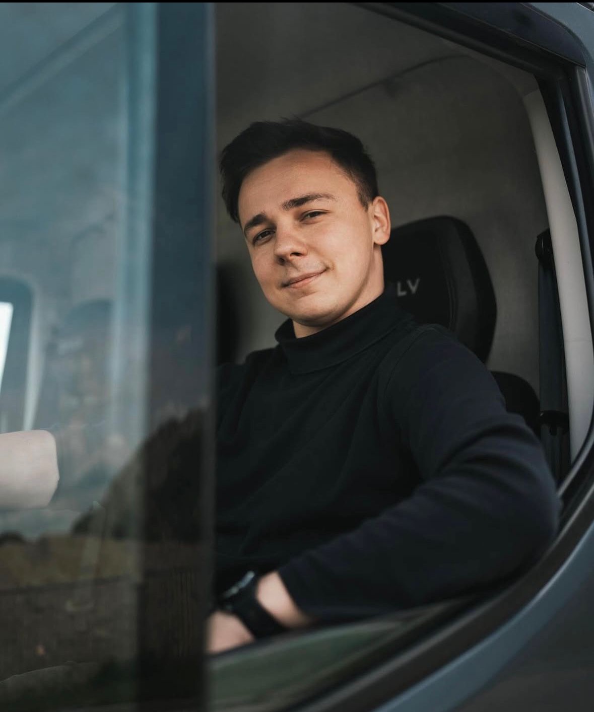 Młody mężczyzna w czarnym golfie siedzi w kabinie nowoczesnego samochodu dostawczego, widoczny przez szybę, z logo 'LV' na zagłówku.