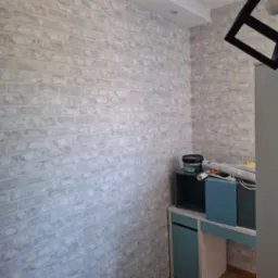 Ściana wykończona tapetą imitującą cegłę w odcieniach szarości. Widoczne akcesoria malarskie i fragment mebli. Czarna lampa w geometrycznym stylu.