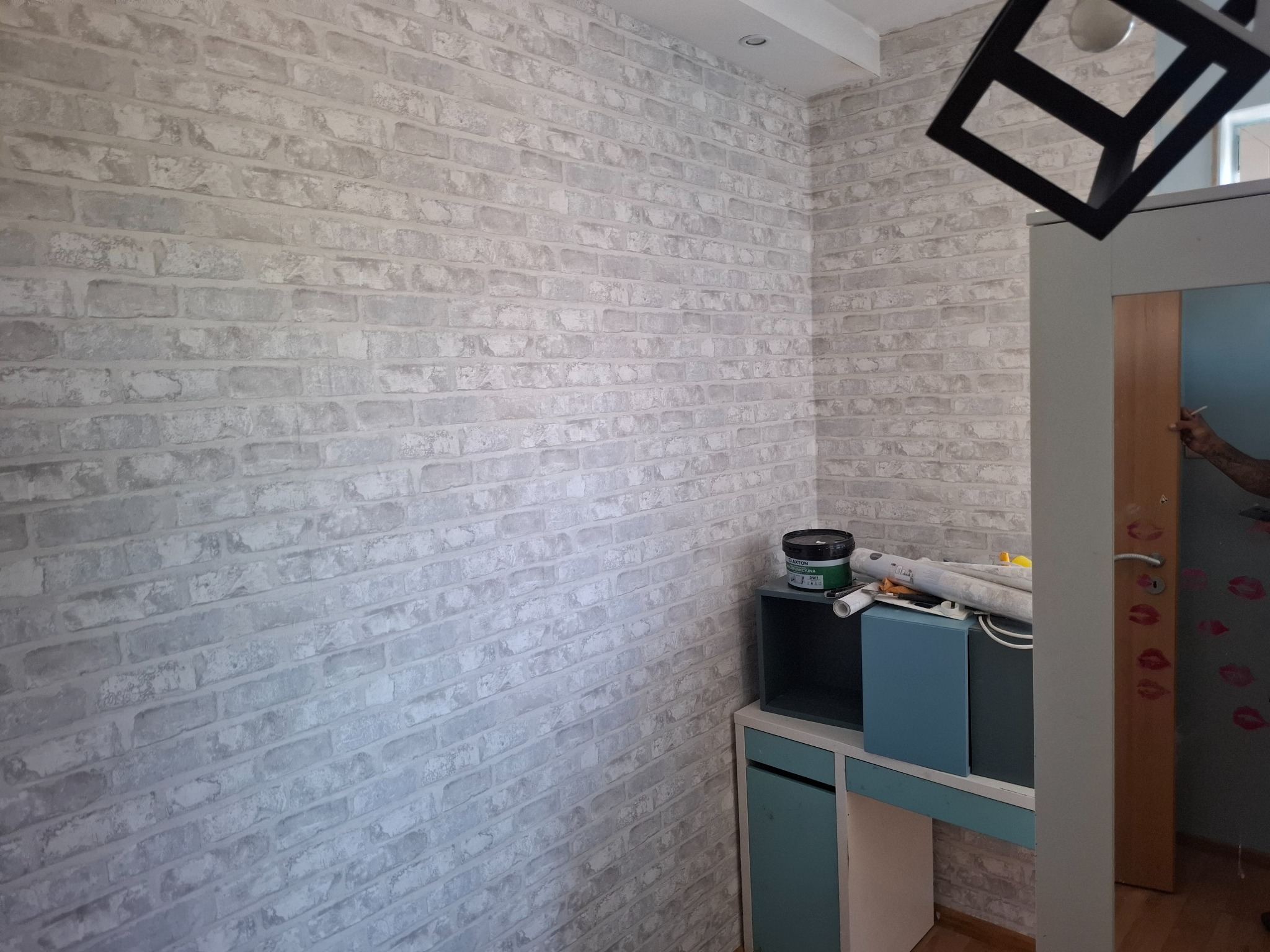 Ściana wykończona tapetą imitującą cegłę w odcieniach szarości. Widoczne akcesoria malarskie i fragment mebli. Czarna lampa w geometrycznym stylu.