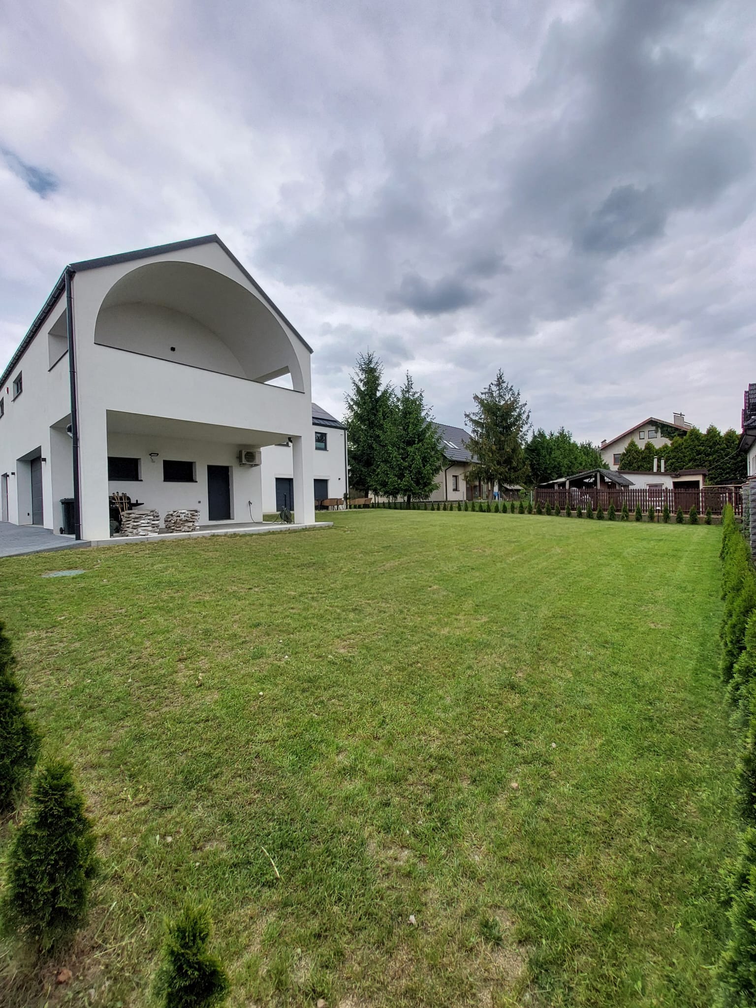 Rozległy trawnik przed nowoczesnym domem z białymi ścianami i łukowym balkonem. W tle widoczne drzewa i ogrodzenie, a na trawniku równo przystrzyżona trawa.