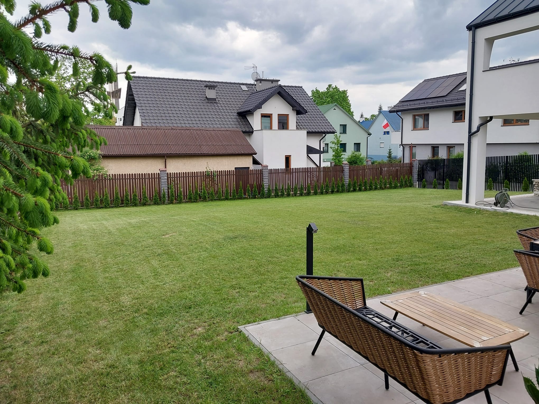 Zadbany trawnik z niską trawą, otoczony tujami i brązowym płotem. W tle zabudowa jednorodzinna z dachami w odcieniach szarości. Na pierwszym planie meble ogrodowe.