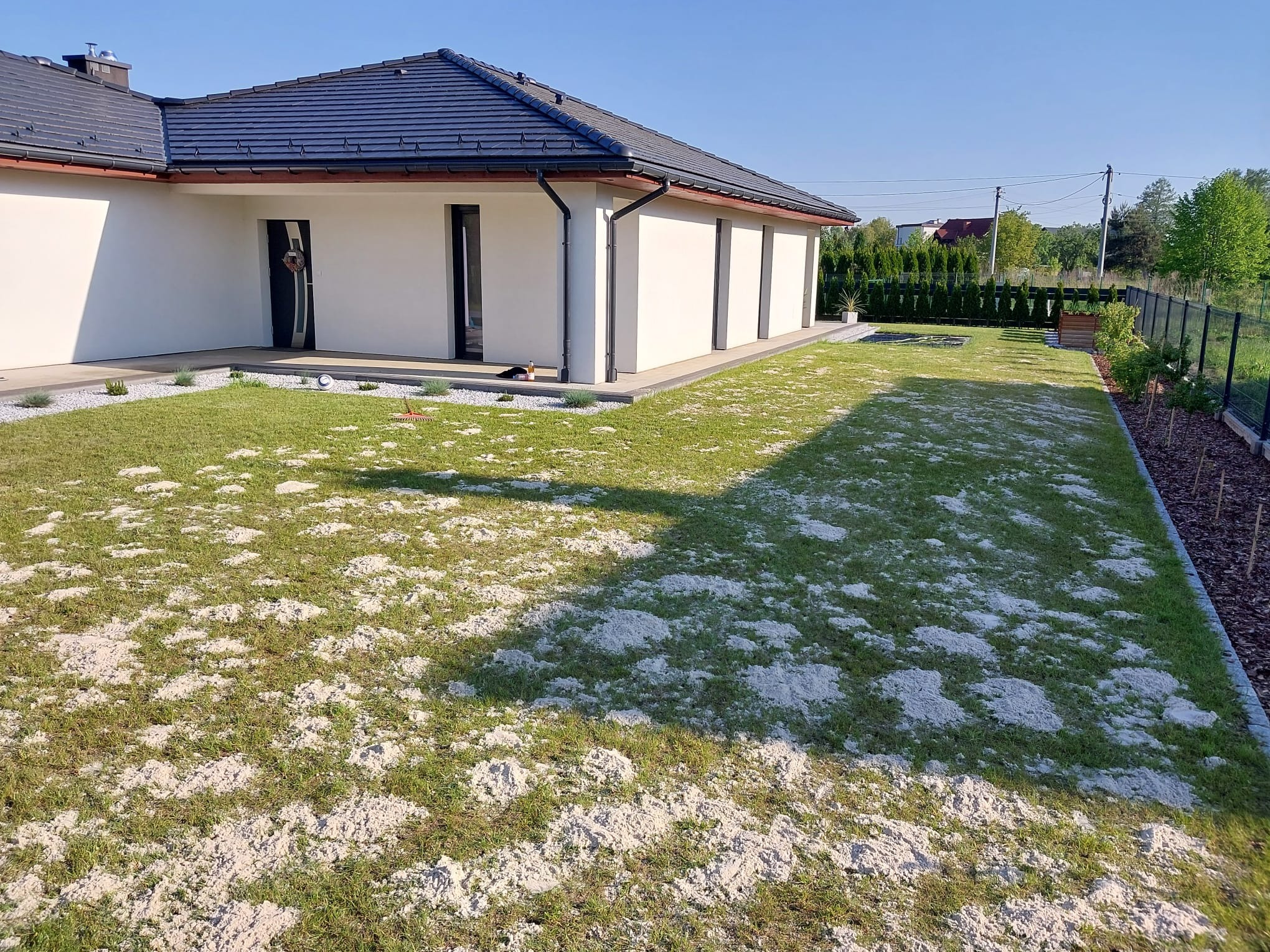 Ogród po zabiegach pielęgnacyjnych z widocznymi kopcami ziemi na trawniku, nowoczesny dom z ciemną dachówką w tle, rabaty z białym kamieniem.