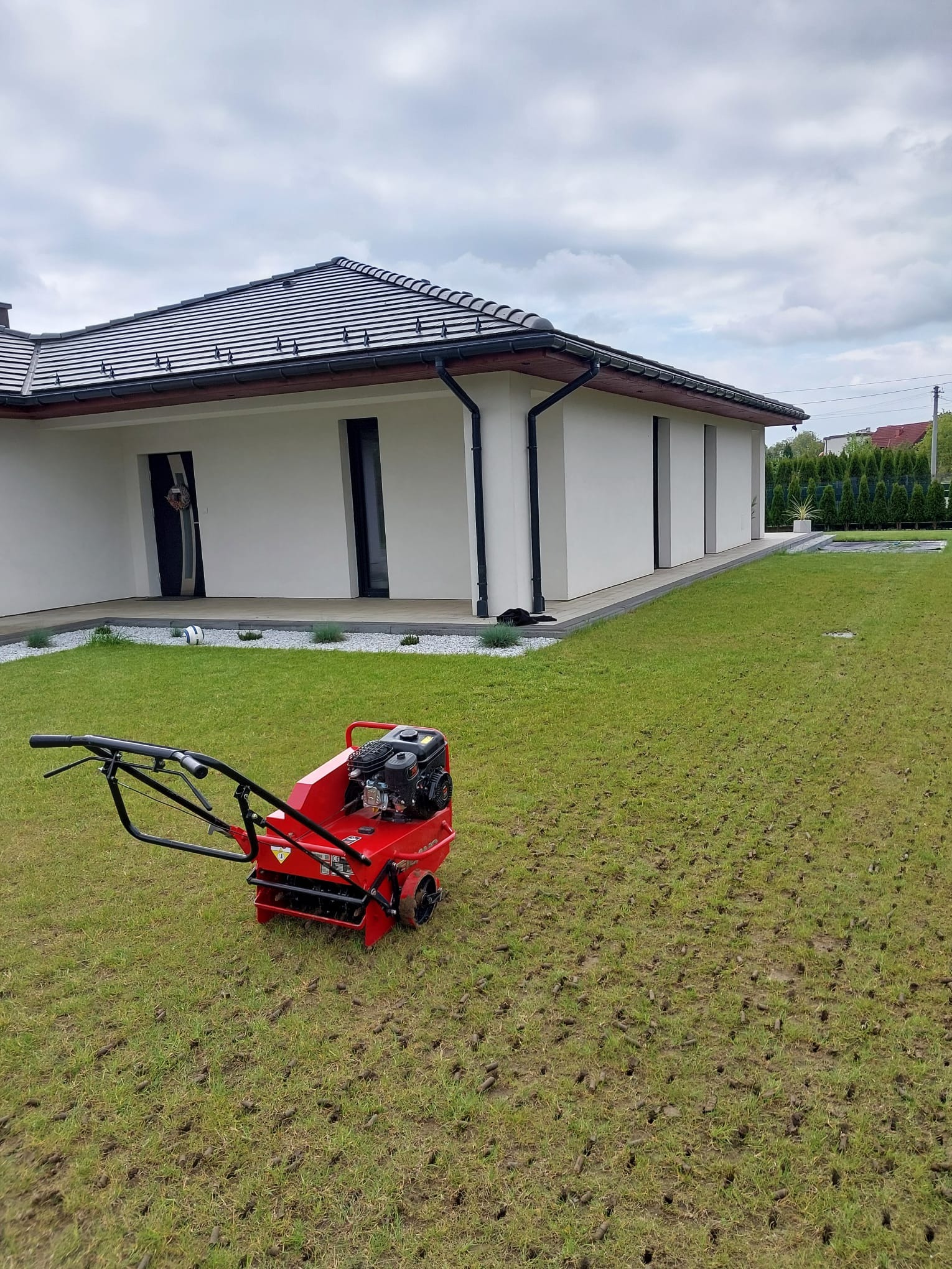 Czerwony aerator na trawniku z widocznymi otworami po aeracji, nowoczesny dom z białymi ścianami w tle, pochmurne niebo.