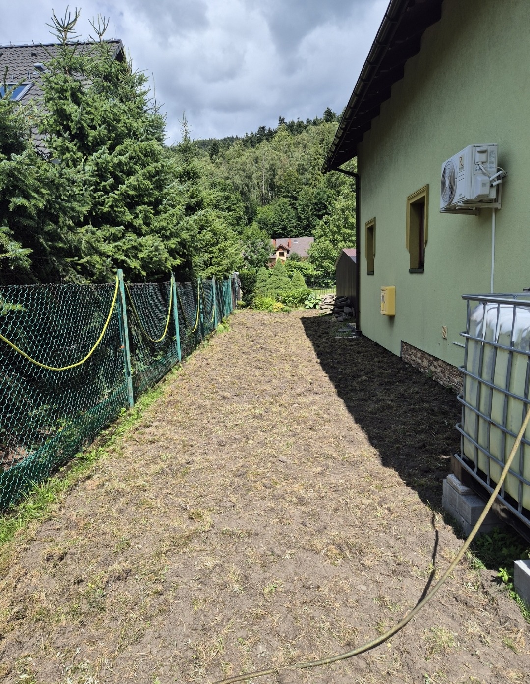 Przygotowany teren pod trawnik z rolki, widoczny system nawadniania. Z tyłu budynek z klimatyzacją i las. Działka obok ogrodzona siatką z żółtym wężem.