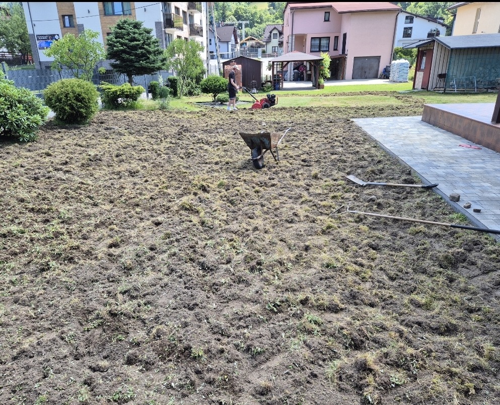 Przygotowany teren pod trawnik z rolki: gleba po wertykulacji, grabie, łopata, taczka i wertykulator w tle. Widok z góry.