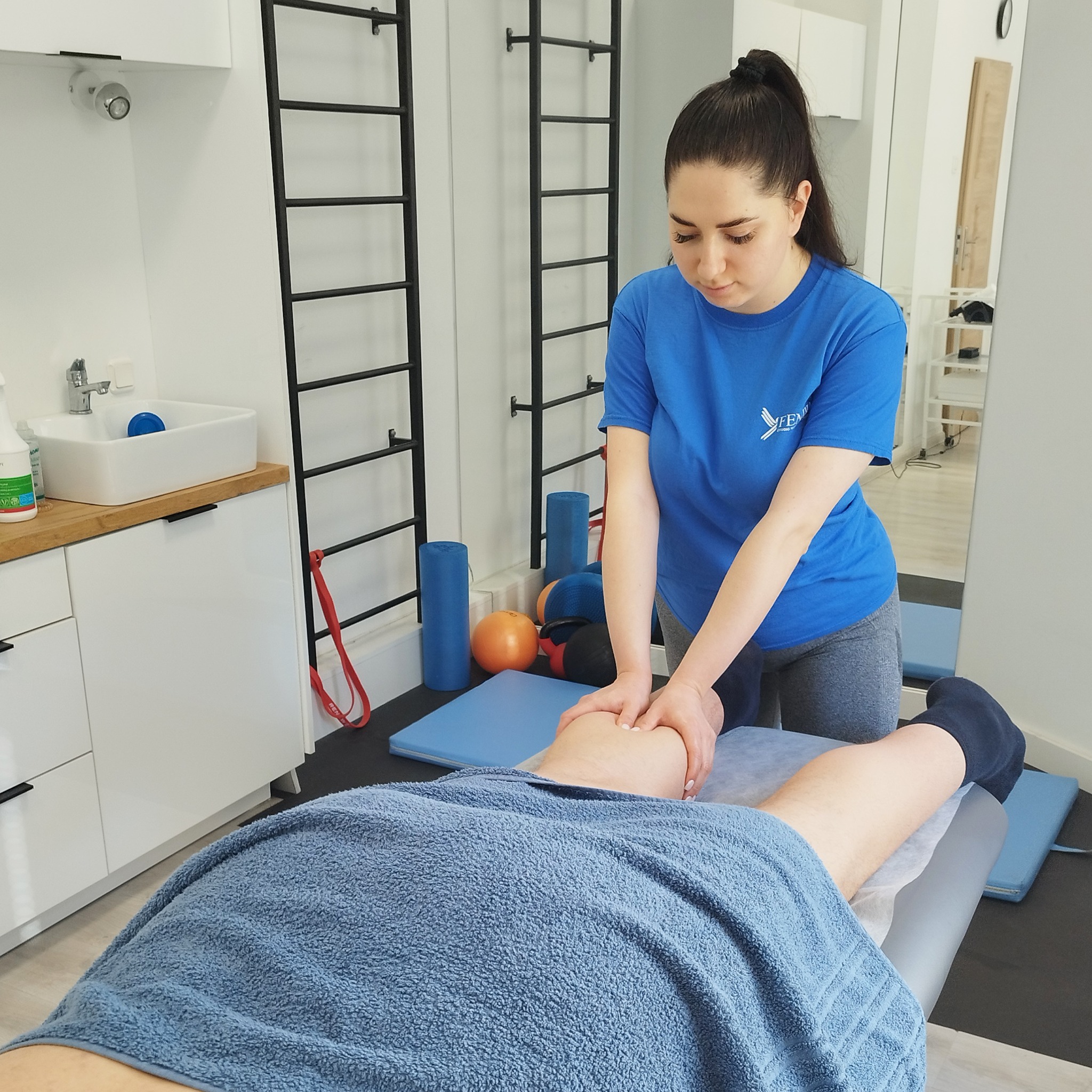 Kobieta w niebieskiej koszulce wykonuje masaż nogi pacjenta leżącego na leżance w gabinecie z drabinkami gimnastycznymi i akcesoriami do ćwiczeń.