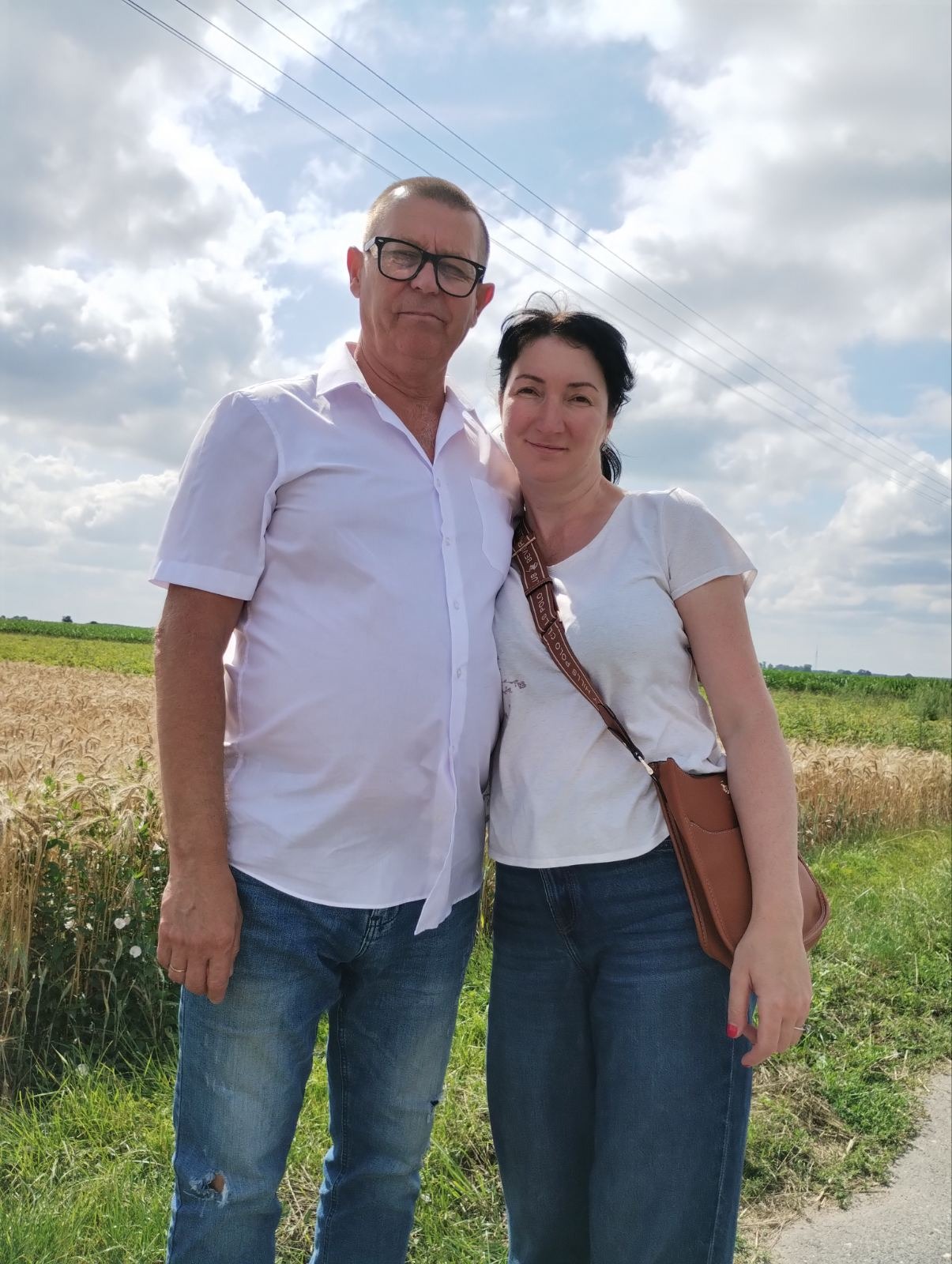 Para w casualowych strojach pozuje na tle pola zboża i błękitnego nieba z chmurami. Mężczyzna w okularach, biała koszula, jeansy z dziurą. Kobieta z torebką przez ramię.