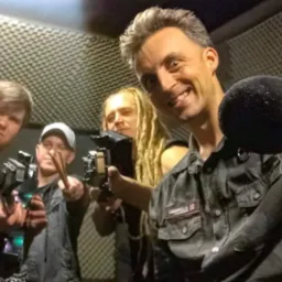 Zespół muzyczny w studiu nagraniowym, mikrofon w dłoni, perkusista z pałeczkami, ścianki akustyczne w tle, uśmiechnięty wokalista, specyficzne oświetlenie.