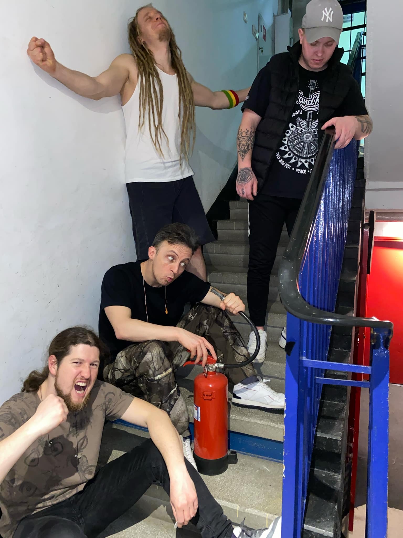 Czterech mężczyzn w różnym wieku pozuje na klatce schodowej w budynku. Jeden z nich trzyma gaśnicę. Ubrani są w casualowe stroje, a ich pozy są dynamiczne i ekspresyjne.