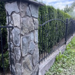 Jacek bla - Kamienny słupek ogrodzeniowy z kutą, metalową balustradą na betonowej podmurówce, na tle zielonych tuj, w pochmurny dzień.