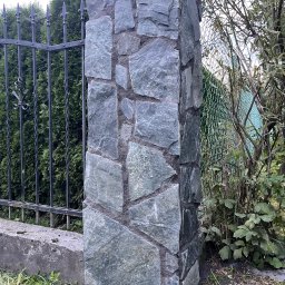 Jacek bla - Kamienna kolumna ogrodzeniowa w odcieniach szarości i zieleni, z widoczną fakturą kamienia, kontrastuje z ciemnym, metalowym ogrodzeniem i zielenią otoczenia.