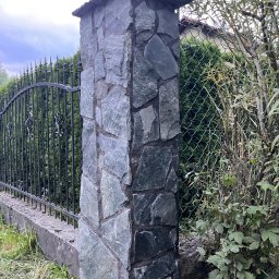 Jacek bla - Kamienny słupek ogrodzeniowy z nieregularnych płyt, przylegający do metalowego ogrodzenia i zieleni. Widoczne detale faktury kamienia i elementy dekoracyjne ogrodzenia.