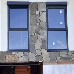 Jacek bla - Dwa okna z roletami, otoczone kamienną elewacją na jasnym budynku. Widoczne fragmenty wnętrza w surowym stanie.