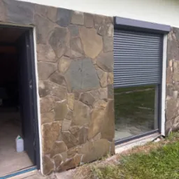 Elewacja z kamienia naturalnego w odcieniach brązu i szarości, okno z roletą zewnętrzną, otwarte drzwi. Wykończenie domu z użyciem kamienia.
