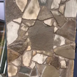 Kamienna elewacja z nieregularnych, brązowo-beżowych płyt, ułożonych na ścianie z wyraźnymi spoinami. Fragment okna i ciemny element konstrukcyjny po bokach.