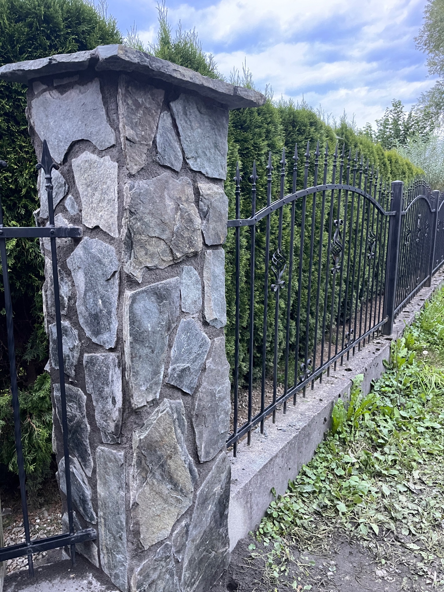 Kamienny słupek ogrodzeniowy z kutą, metalową balustradą na betonowej podmurówce, na tle zielonych tuj, w pochmurny dzień.