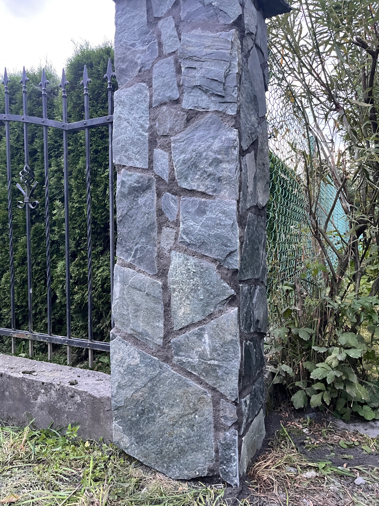 Kamienna kolumna ogrodzeniowa w odcieniach szarości i zieleni, z widoczną fakturą kamienia, kontrastuje z ciemnym, metalowym ogrodzeniem i zielenią otoczenia.