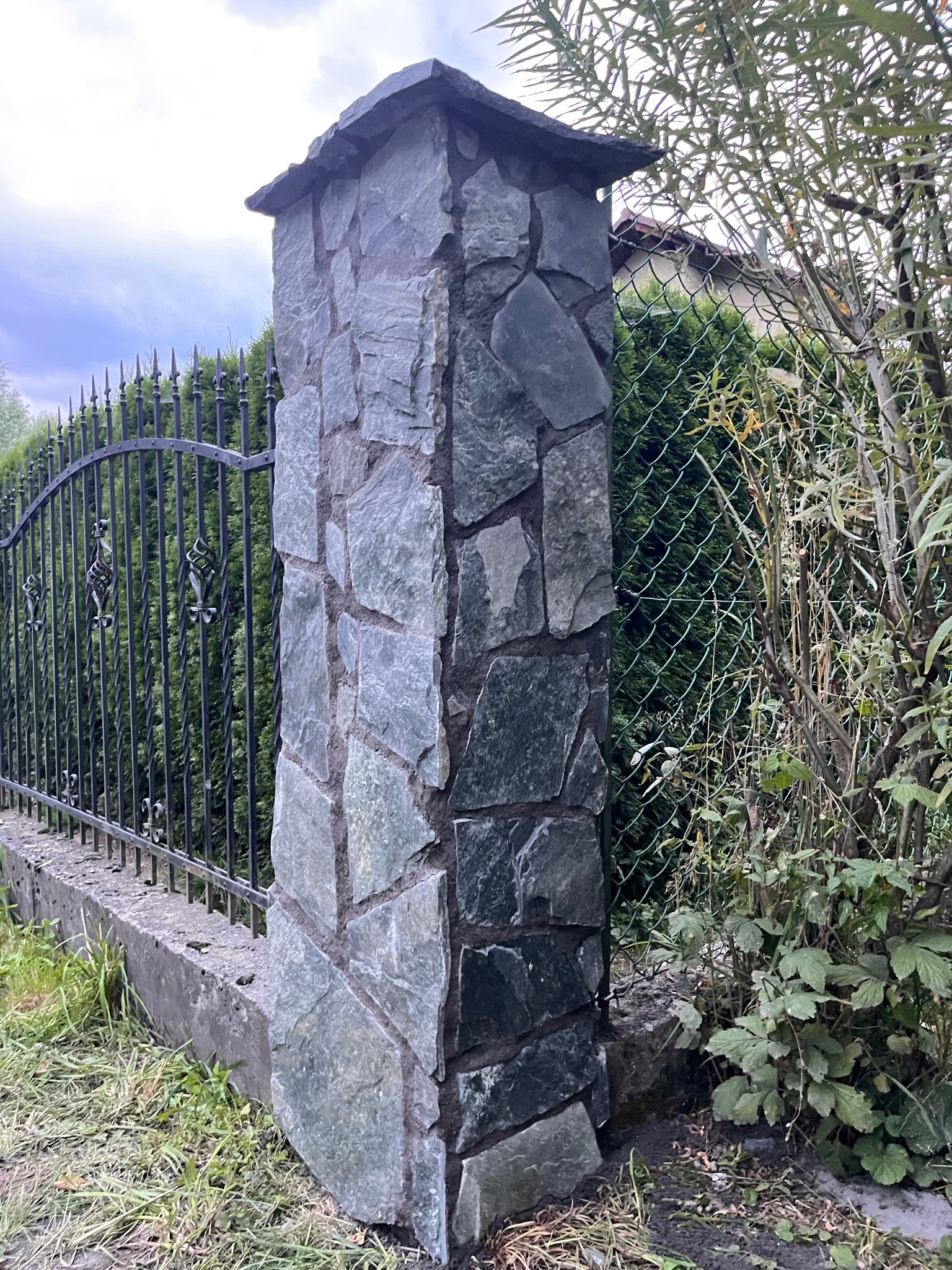 Kamienny słupek ogrodzeniowy z nieregularnych płyt, przylegający do metalowego ogrodzenia i zieleni. Widoczne detale faktury kamienia i elementy dekoracyjne ogrodzenia.