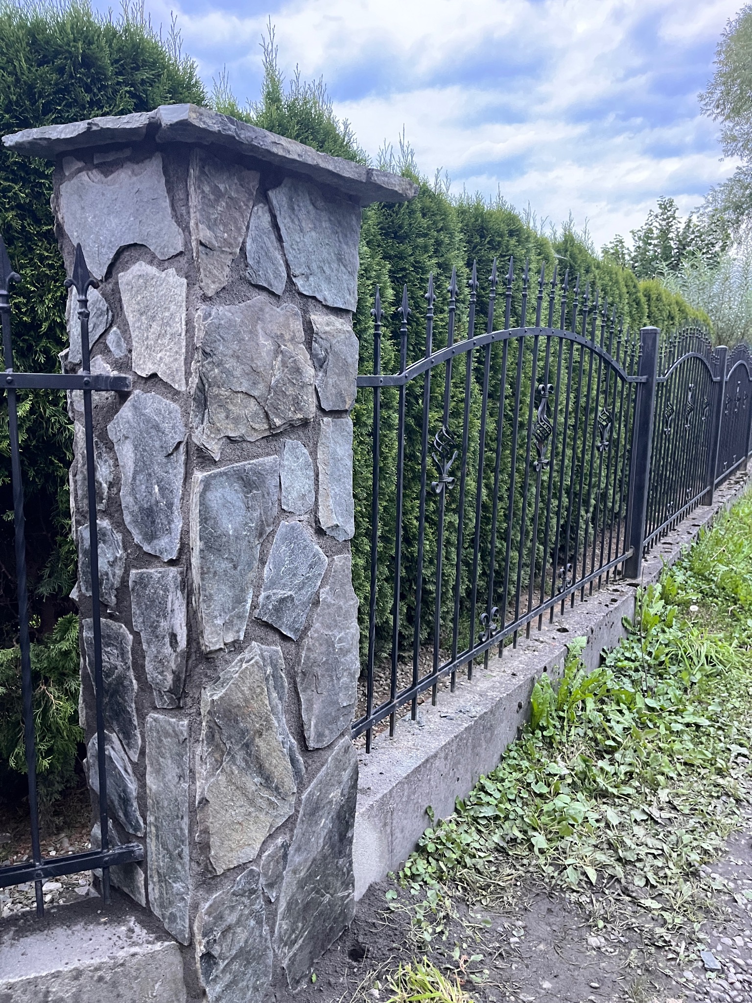 Kamienne słupki ogrodzeniowe z kutym, metalowym ogrodzeniem na tle zielonych krzewów. Detal wykończenia ogrodu, solidne wykonanie.