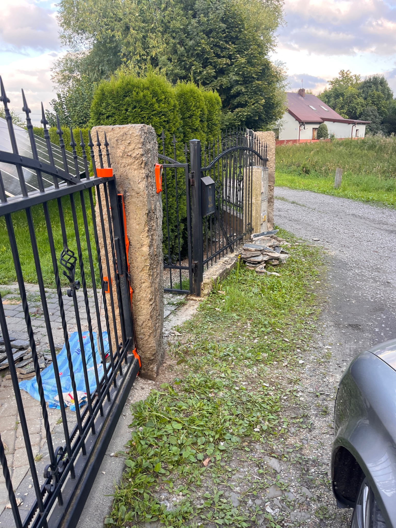 Montaż czarnej, metalowej bramy z kamiennymi słupkami. Widoczne narzędzia i fragment auta. Tło: dom i zieleń. Kąt ujęcia z boku.