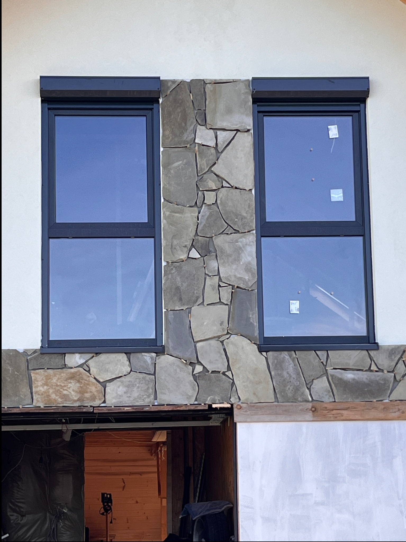 Dwa okna z roletami, otoczone kamienną elewacją na jasnym budynku. Widoczne fragmenty wnętrza w surowym stanie.