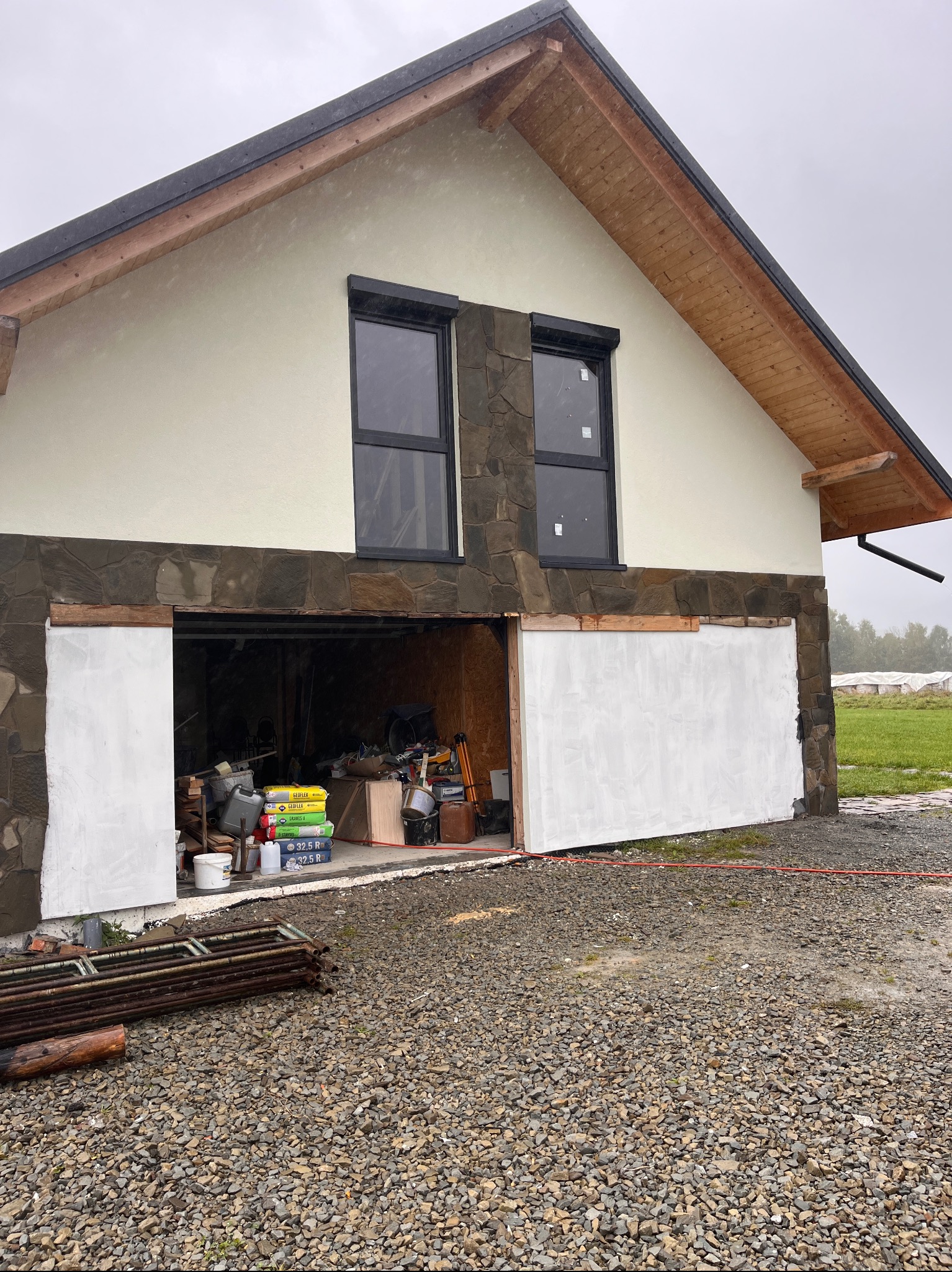 Budynek z elewacją z kamienia i oknami z roletami zewnętrznymi. Częściowo otynkowana ściana garażu z widocznymi narzędziami i materiałami budowlanymi w środku.