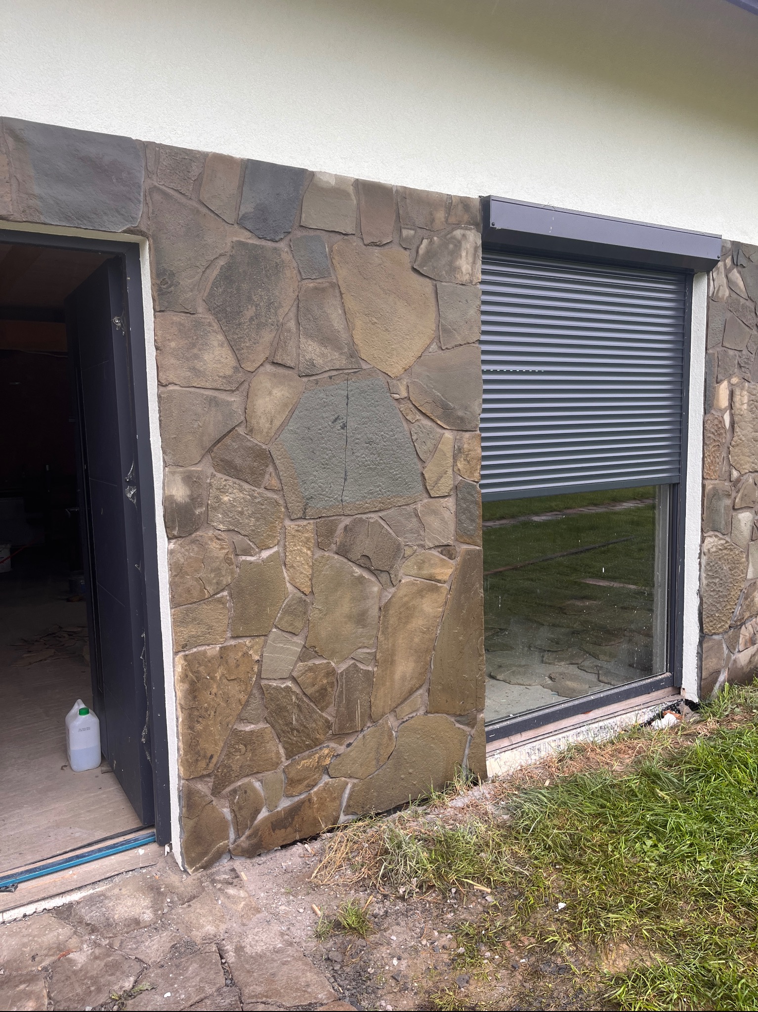 Elewacja z kamienia naturalnego w odcieniach brązu i szarości, okno z roletą zewnętrzną, otwarte drzwi. Wykończenie domu z użyciem kamienia.