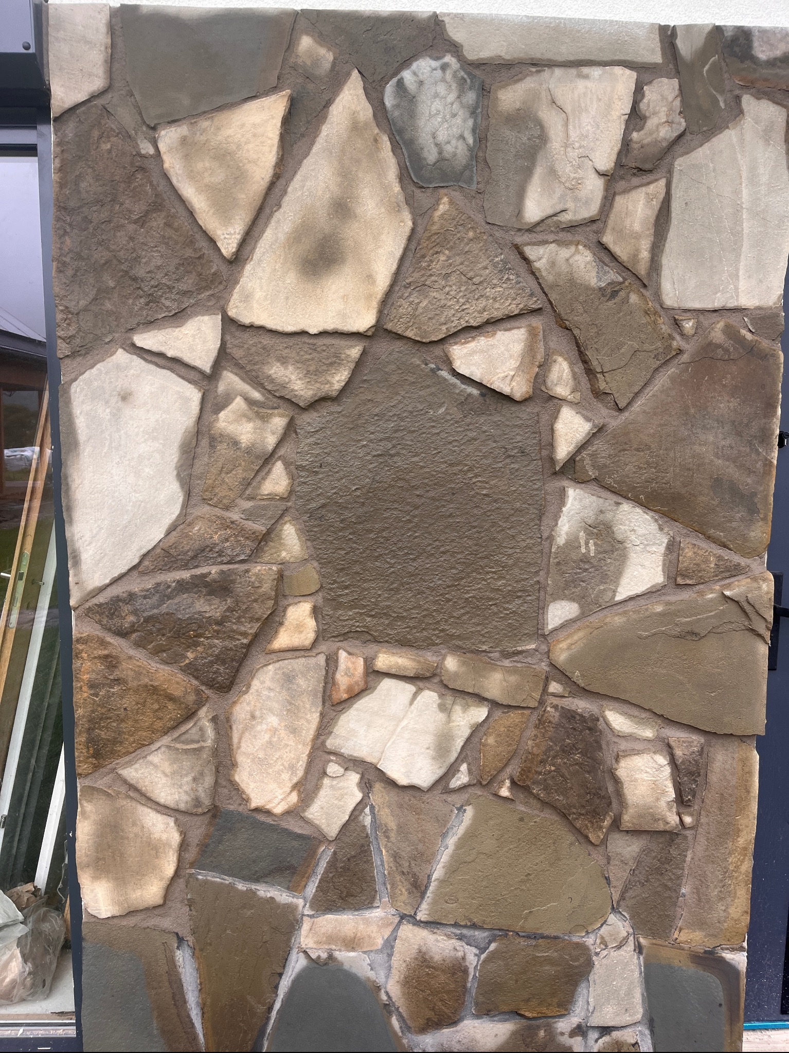 Kamienna elewacja z nieregularnych, brązowo-beżowych płyt, ułożonych na ścianie z wyraźnymi spoinami. Fragment okna i ciemny element konstrukcyjny po bokach.