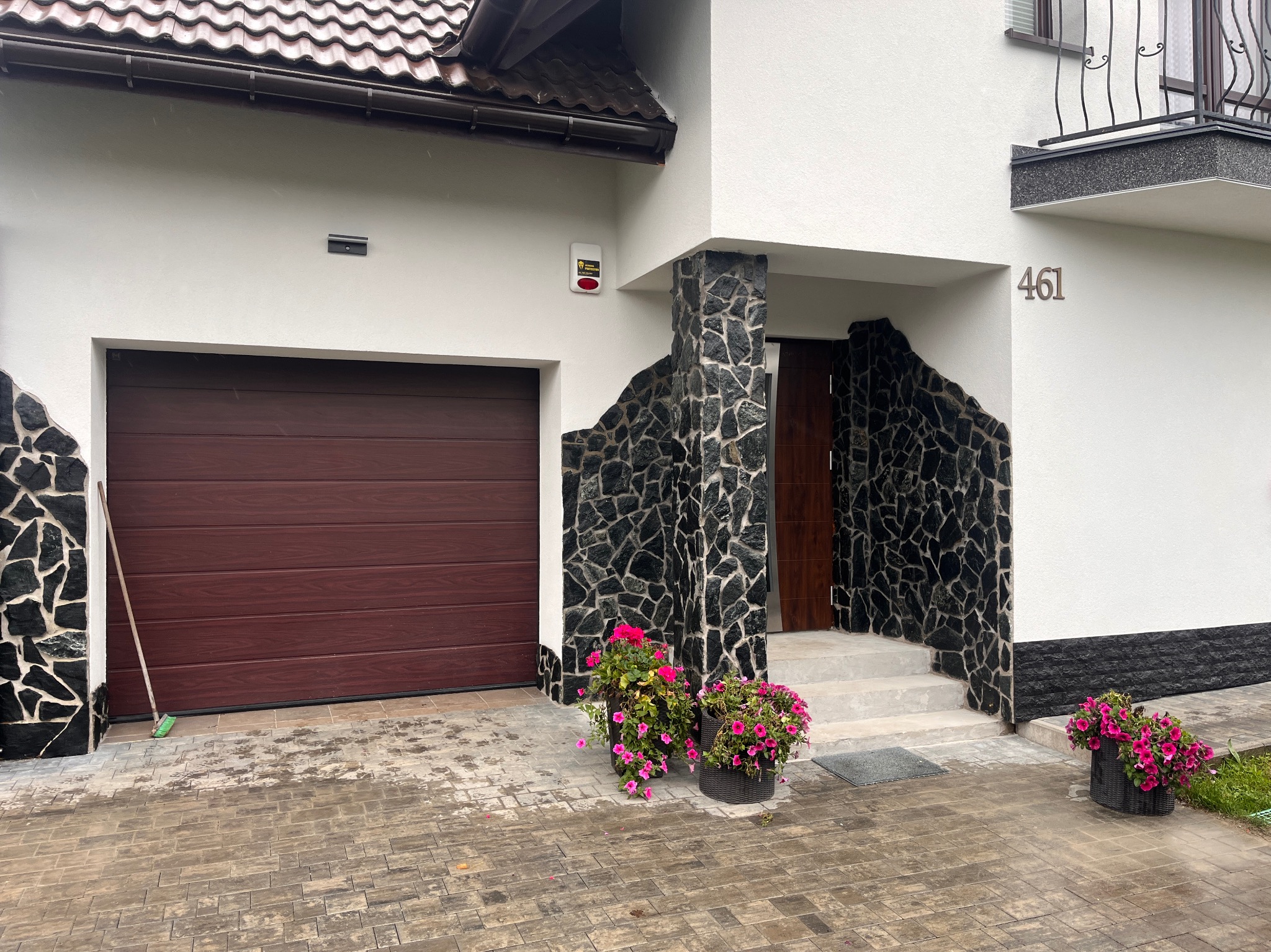 Dom z garażem i wejściem wykończonym kamieniem, numer domu 461. Brukowany podjazd, kwiaty w donicach. Całość utrzymana w stonowanej kolorystyce.