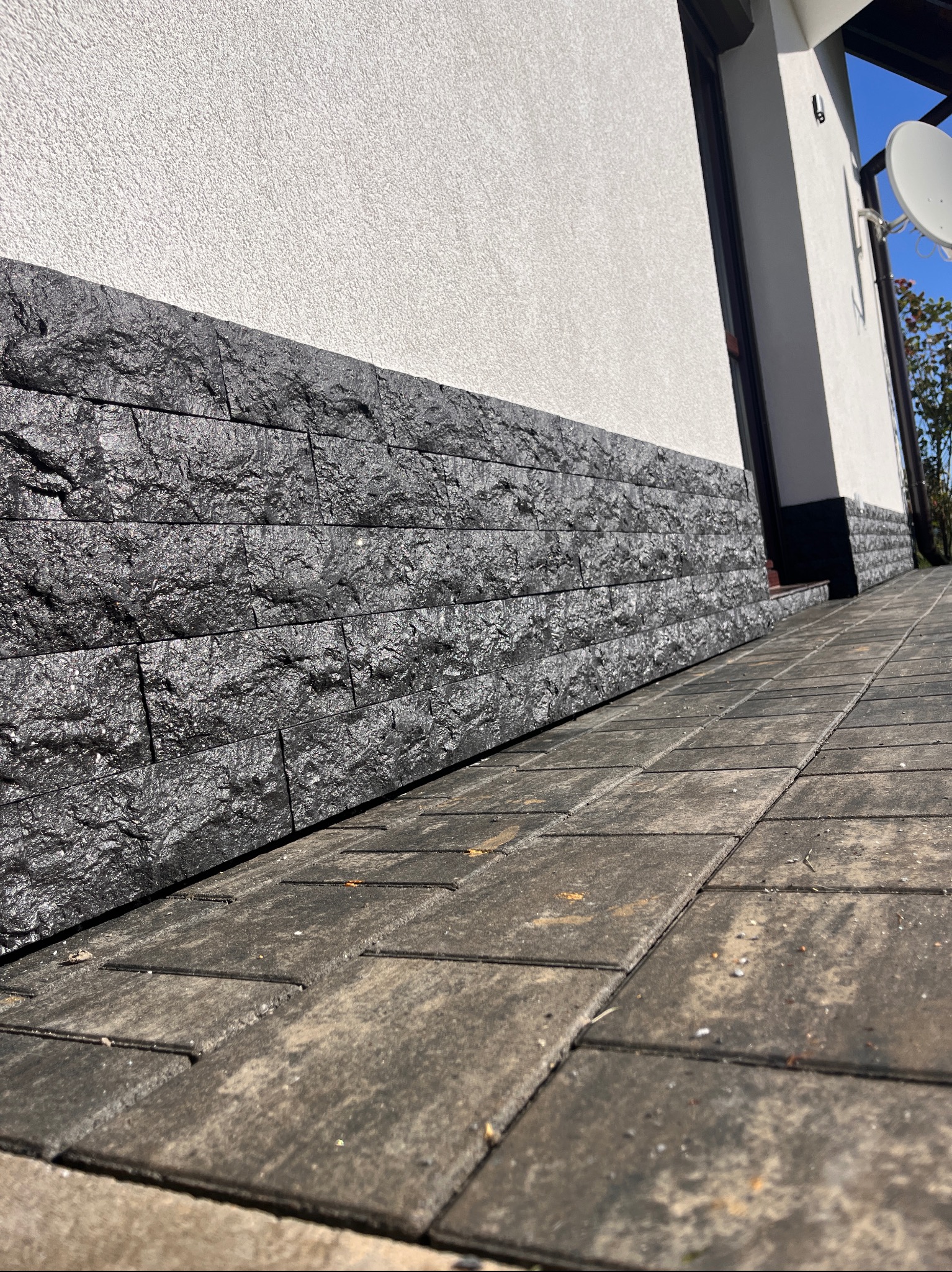 Elewacja domu z czarnym kamieniem dekoracyjnym i szarą kostką brukową. Widoczny fragment ściany i fragment dachu budynku z anteną satelitarną.